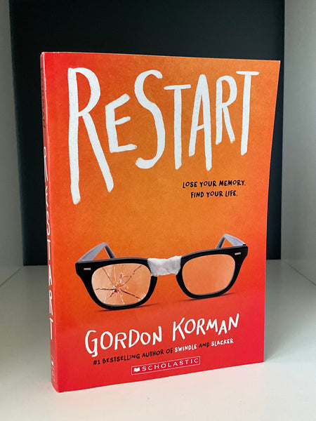 Book cover of Restart by Gordon Korman featuring broken black glasses taped at the bridge on an orange-to-red gradient background, with the tagline ‘Lose your memory. Find your life.’