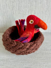 Theo the felted red parrot, nestled in a hand-knit brown wool nest—Formation Flock character from Wonder & Wisdom Books™, photographed on a textured white background.