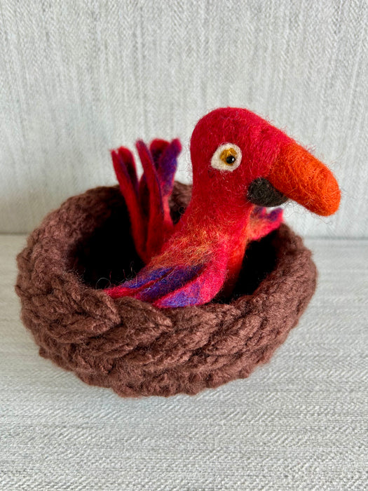 Theo the felted red parrot, nestled in a hand-knit brown wool nest—Formation Flock character from Wonder & Wisdom Books™, photographed on a textured white background.