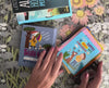 Hands flipping through the What’s for Dinner? Truth Cards deck, showing various illustrated cards with uplifting messages. The Truth Cards box and other books are visible on a floral tablecloth.