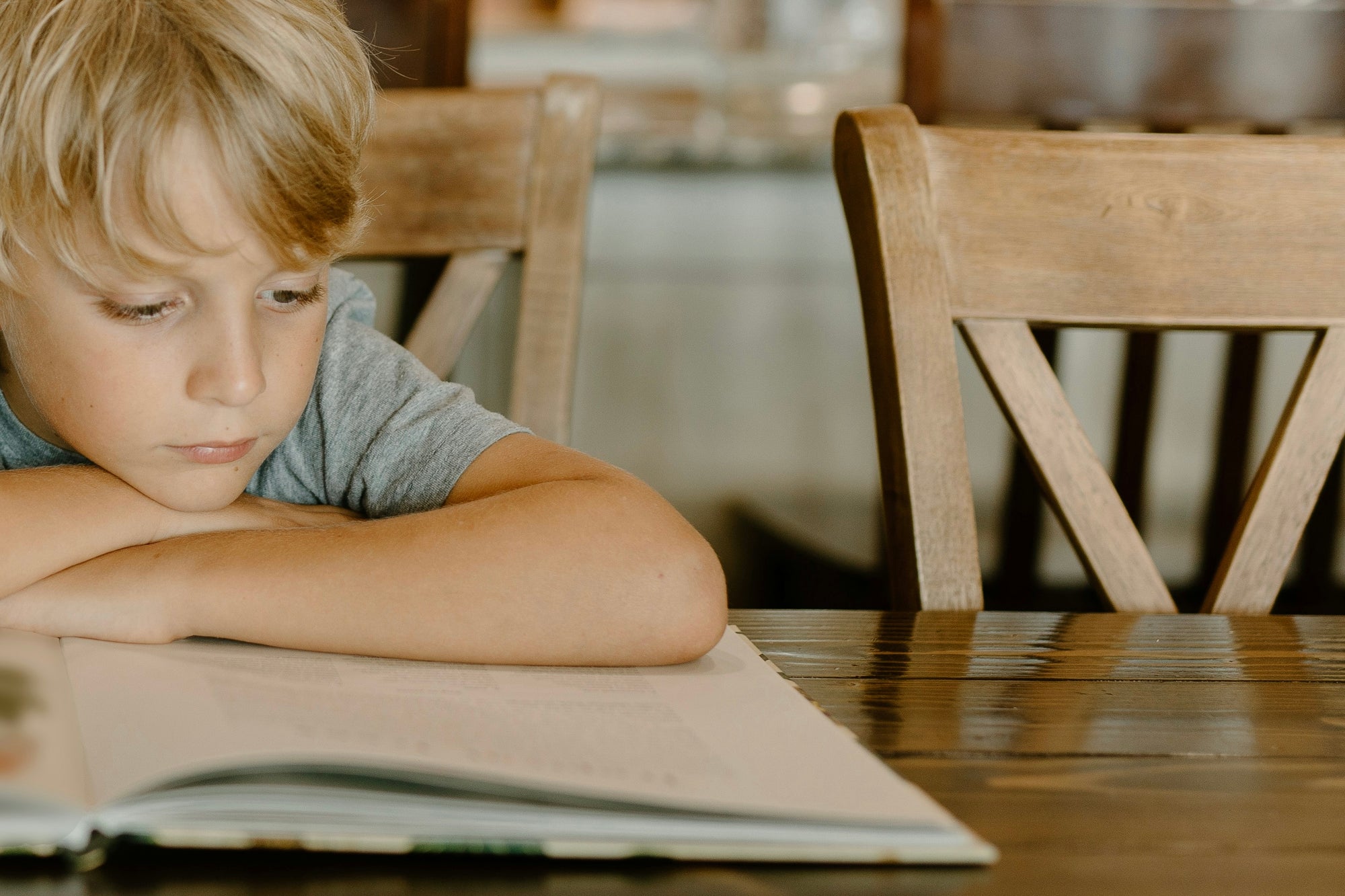 Child resting on folded arms while reading an open book at a wooden table in warm natural light
